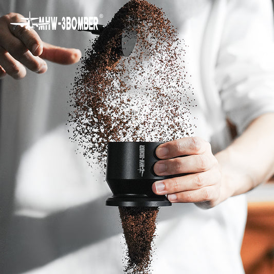 A person in a white shirt holds an MHW-3BOMBER Espresso Coffee Powder Dosing Funnel as ground coffee beans are poured in with flair, spilling over. The scene captures the artistry and precision loved by home enthusiasts and baristas alike.
