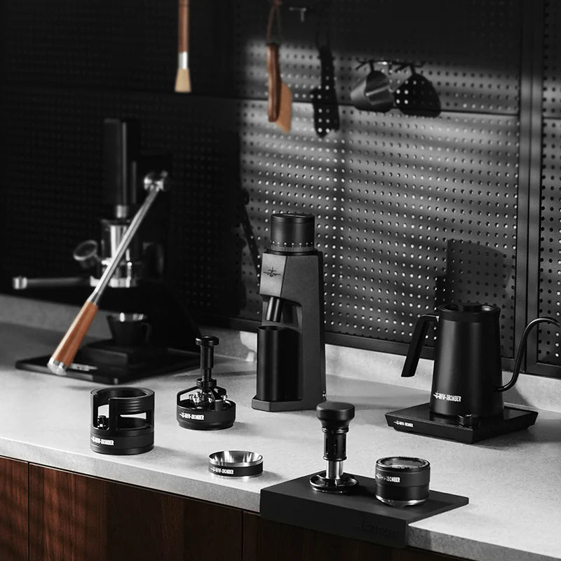 MHW-3BOMBER black coffee tools: distributor, leveler, kettle, grinder, tamper, and accessories on counter.