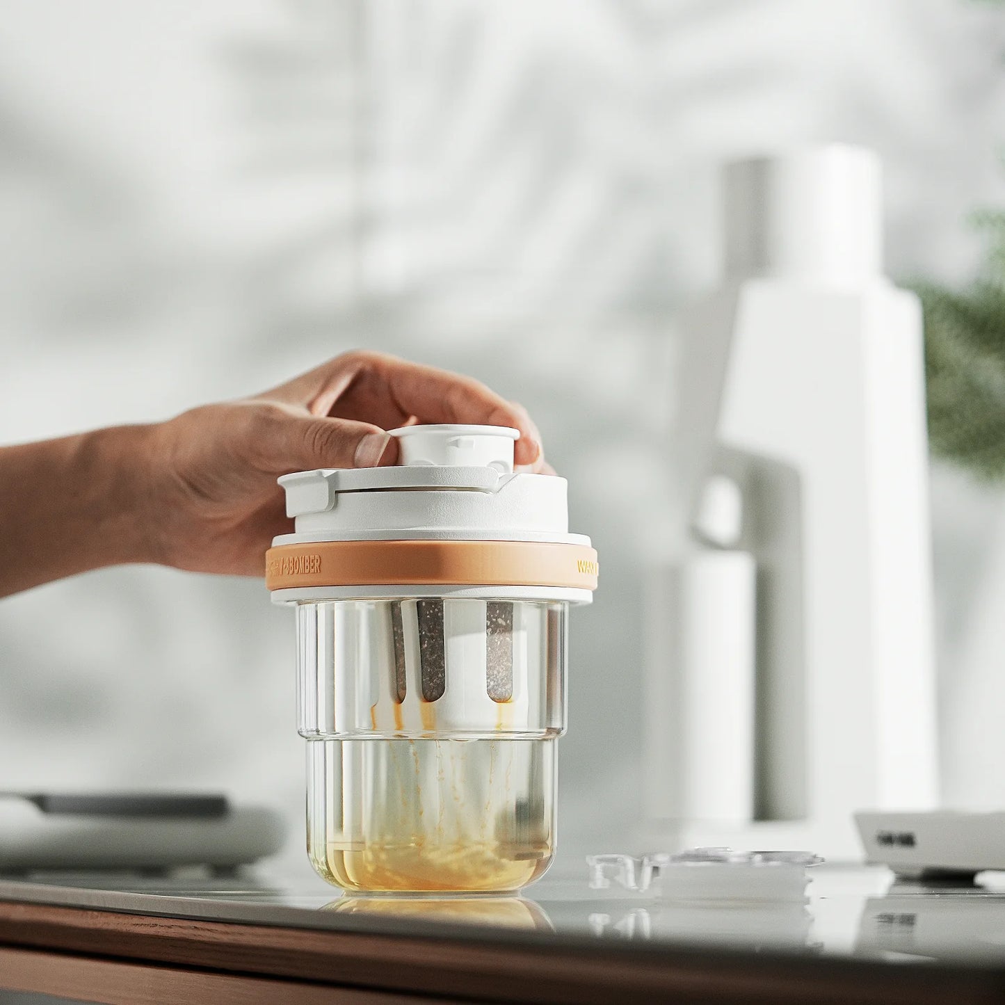 MHW-3BOMBER cold brew cup on counter. Clear with orange accent, white lid. Portable, leak-proof design. In use.