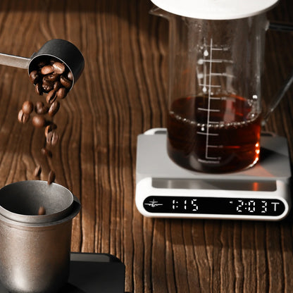 MHW-3BOMBER smart coffee scale with timer & beaker. Coffee beans falling. On a wooden surface.