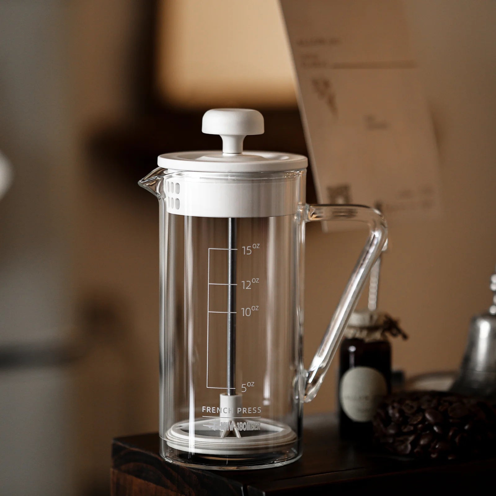 The MHW-3BOMBER Gimme French Press, with a clear glass and classic design featuring a white lid and measurement lines up to 15 oz., rests on wood. A premium stainless steel filter ensures smooth brewing. In the blurred background are a small bottle and stacked papers or cards.