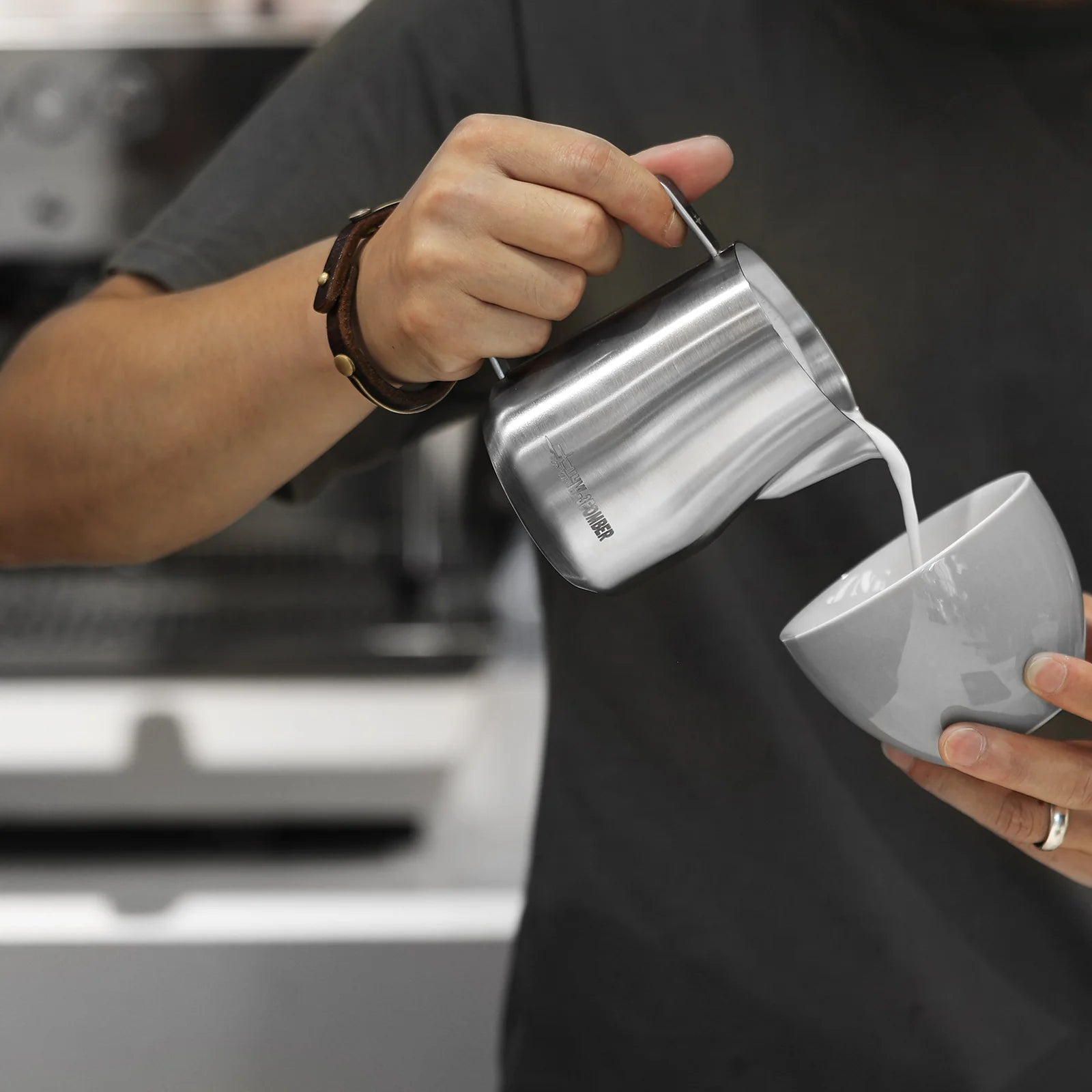 Wearing a gray shirt and a brown watch, someone expertly pours milk from an MHW-3BOMBER Turbo Milk Pitcher Glossy into a white cup, crafting delicate latte art. A softly blurred coffee machine is visible in the background.
