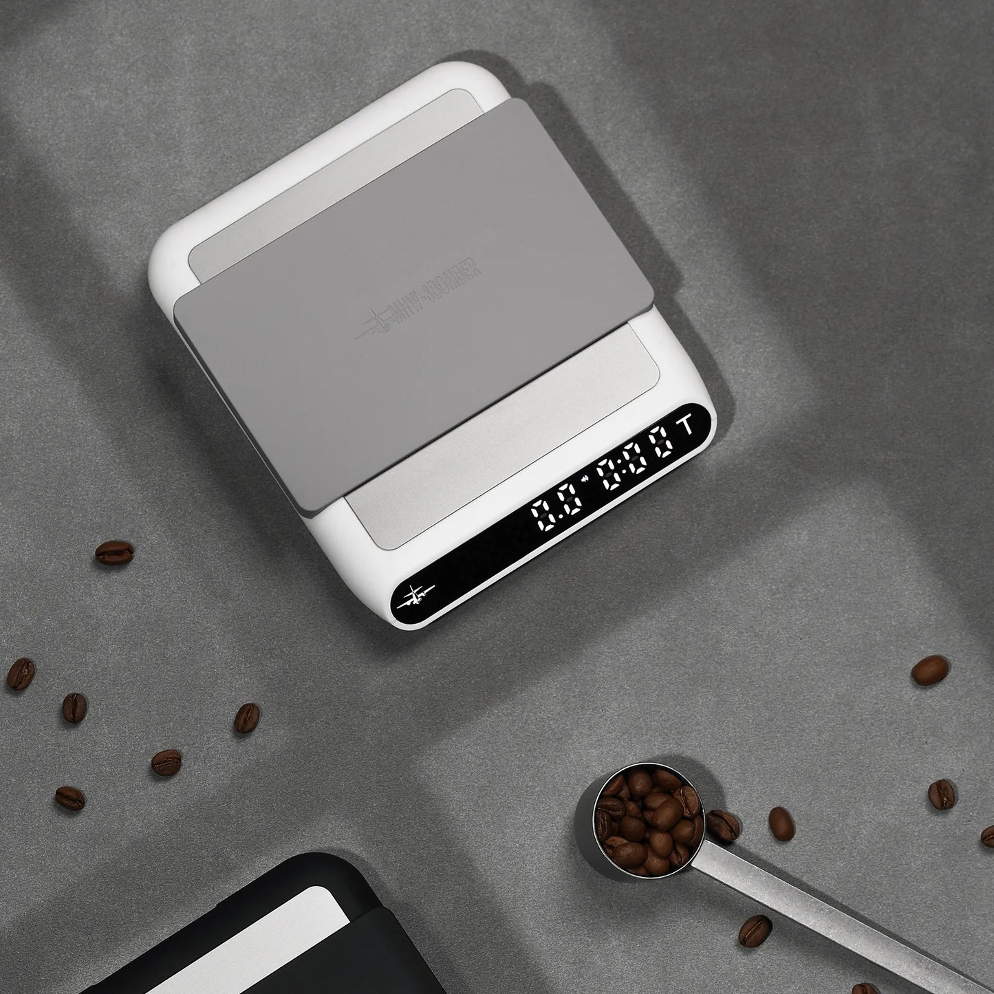 MHW-3BOMBER smart coffee scale. White with gray pad. Digital display. On counter with coffee beans. 