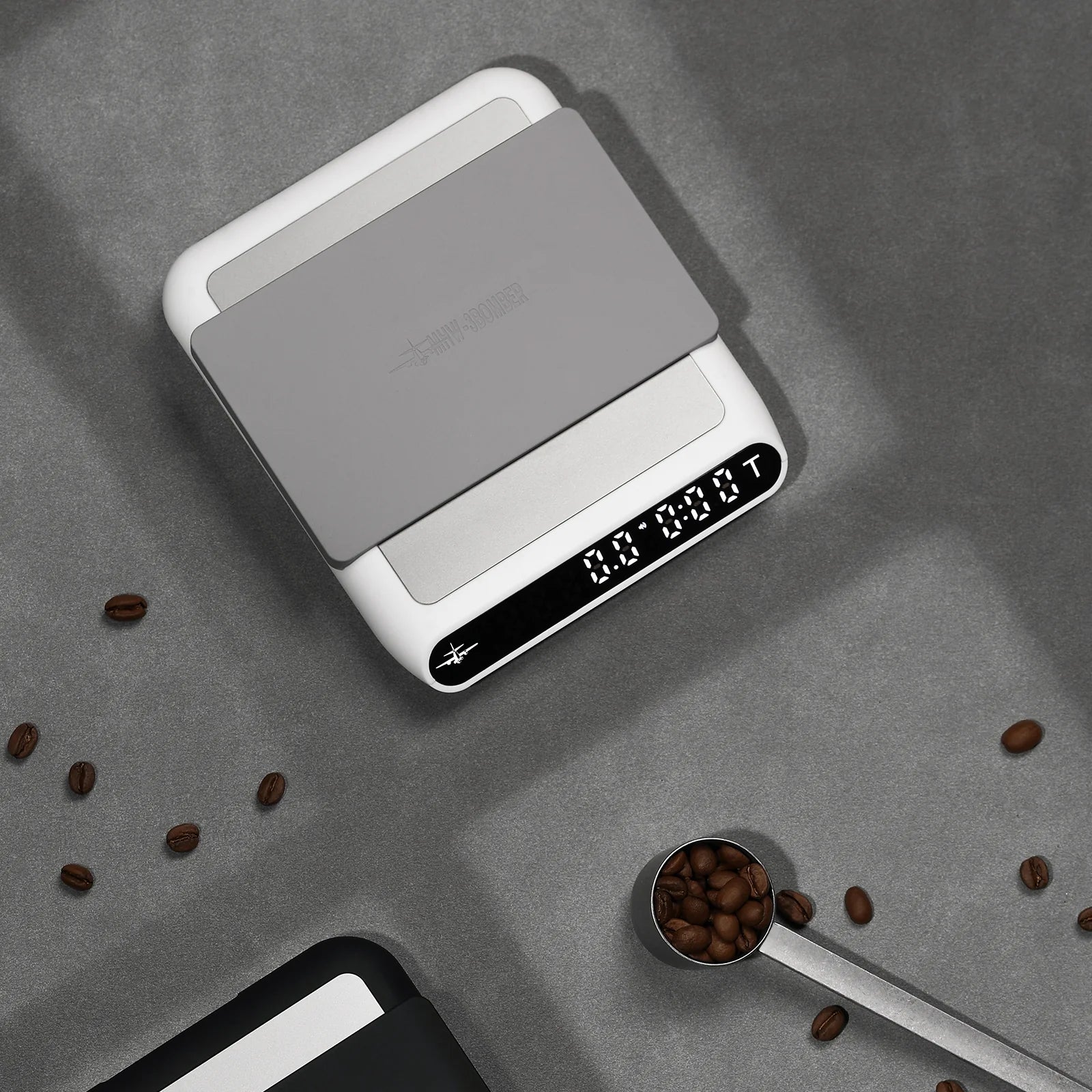 MHW-3BOMBER smart coffee scale. White with gray pad. Digital display. On counter with coffee beans. 