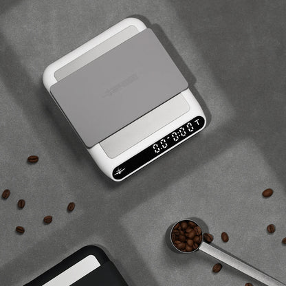 MHW-3BOMBER smart coffee scale. White with gray pad. Digital display. On counter with coffee beans. 