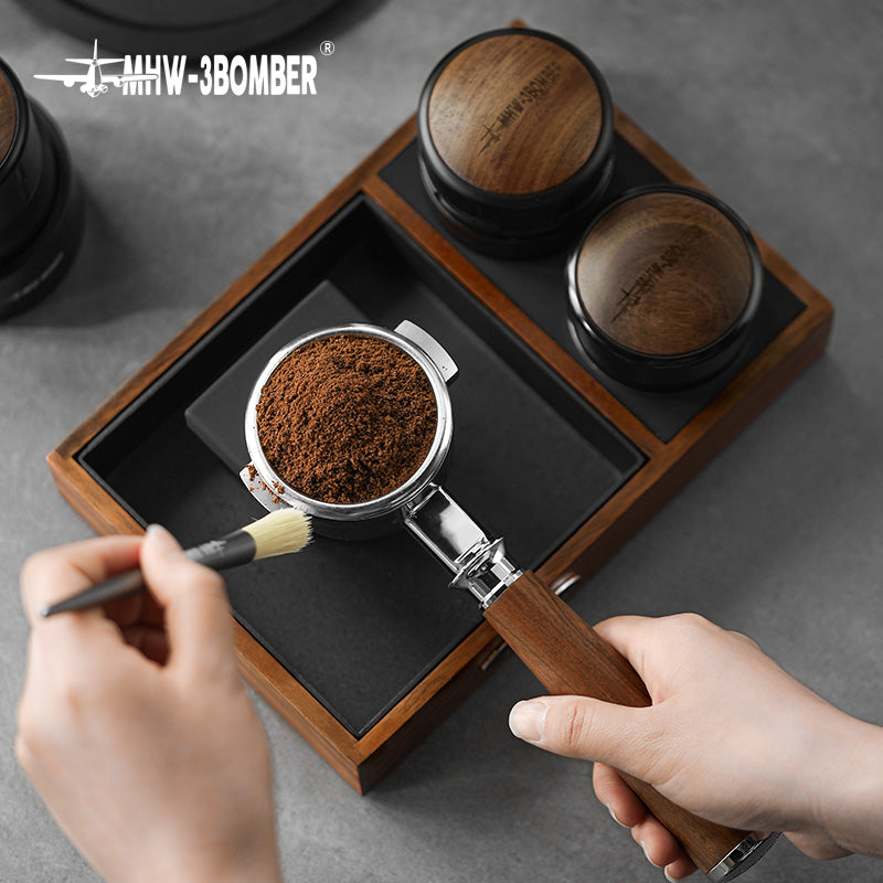 MHW-3BOMBER espresso tamping station with portafilter, tamper, wood base and knock box. Coffee grinds visible.