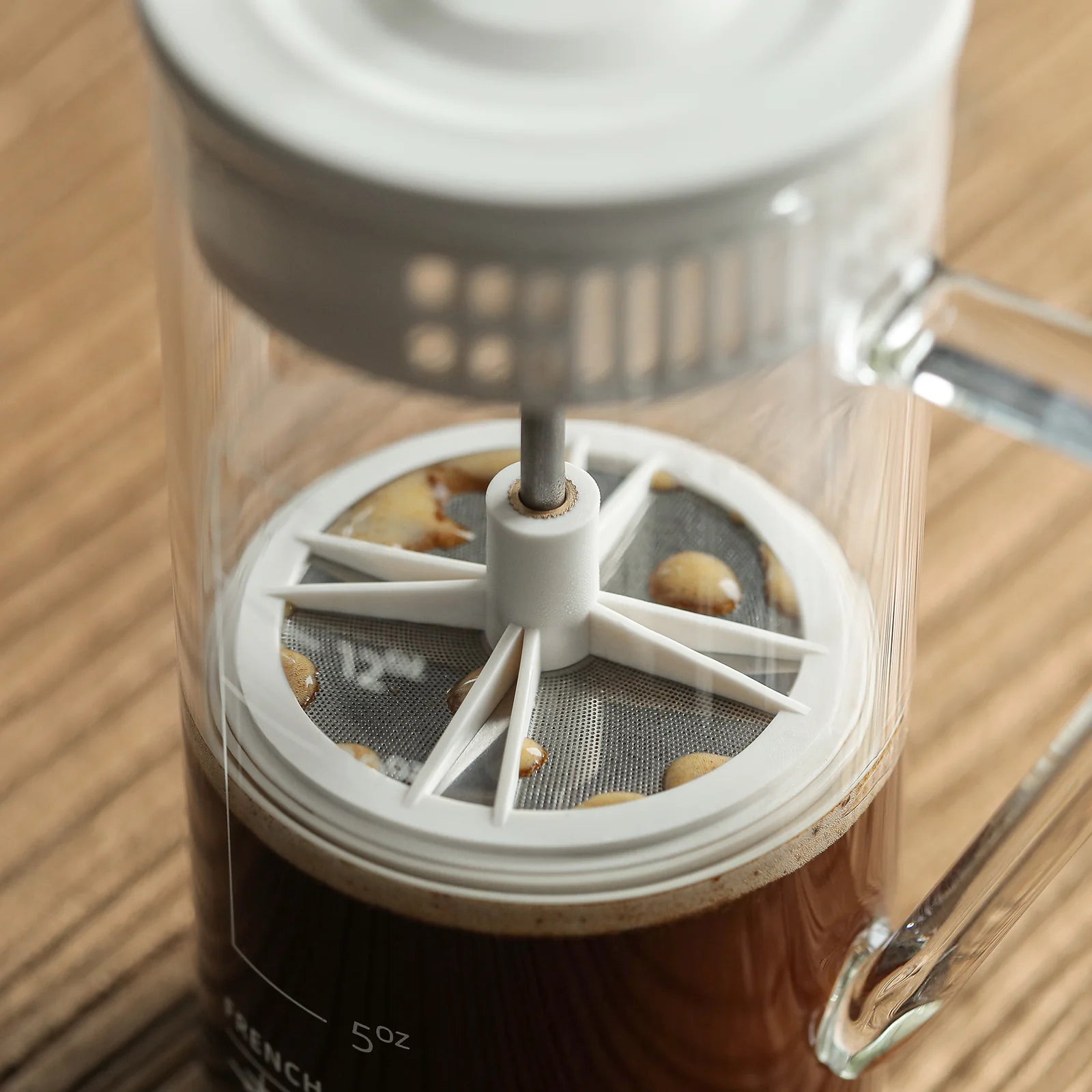 A close-up of the MHW-3BOMBER Gimme French Press reveals its stainless steel filter immersed in dark coffee, showing bubbles on the surface. The heat-resistant glass and metal components are clearly visible, set on a wooden surface.