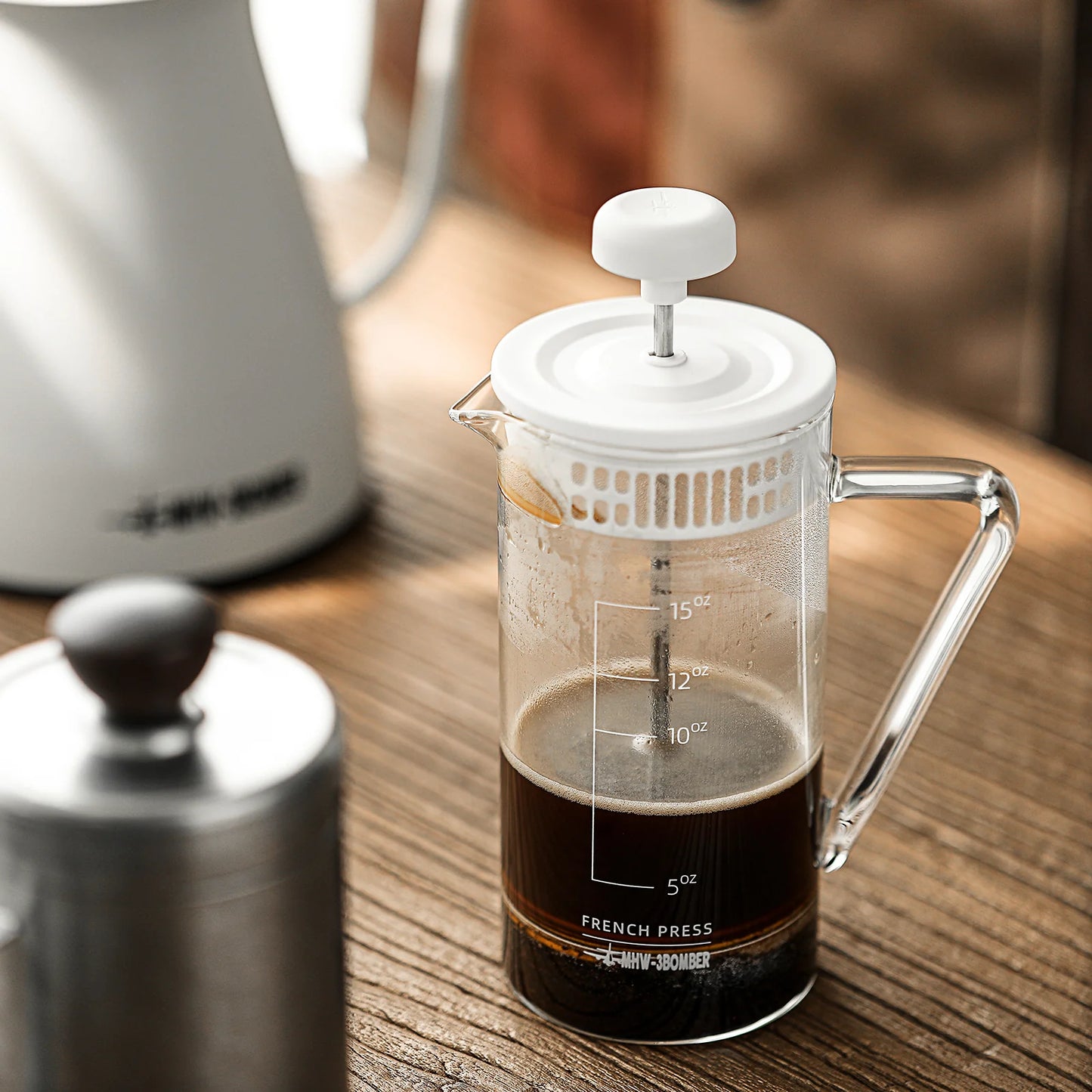 The MHW-3BOMBER Gimme French Press, featuring a sleek white lid and stainless steel filter, holds dark coffee in its heat-resistant glass body with measurement markings. It sits on a wooden table next to a metal pot and a white kettle.