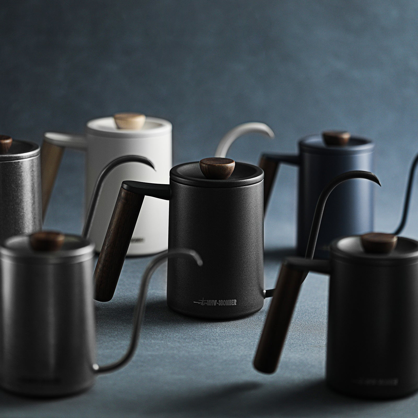 The MHW-3BOMBER Planet Hand Brewing Kettle collection, featuring matte finishes and wooden handles, is perfectly suited for brewing. Displayed on a dark textured surface, these kettles in black, gray, and white are scattered elegantly to create a sleek and modern look.
