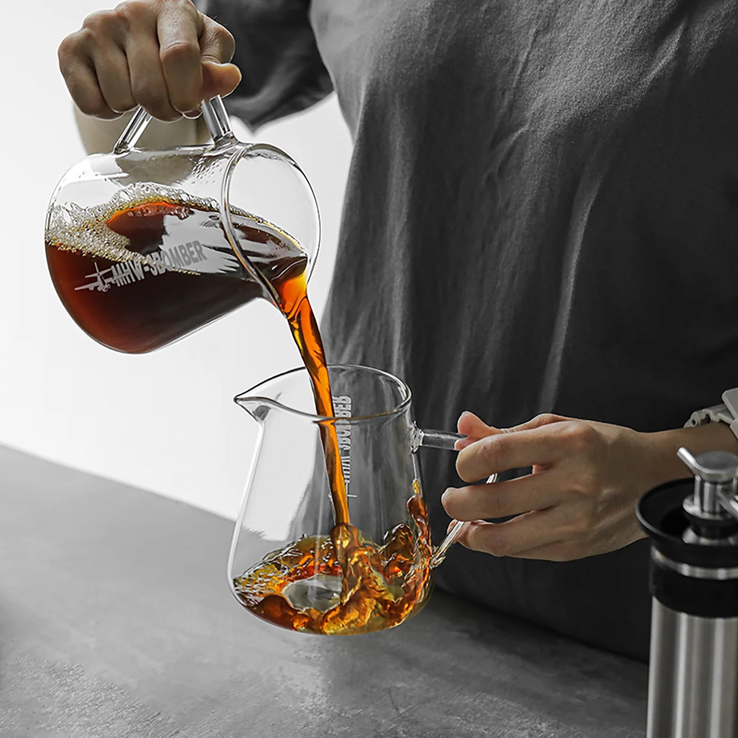 A person in a dark shirt effortlessly pours coffee from a borosilicate glass mug into an MHW-3BOMBER Elf Coffee Server on the gray countertop. A metal coffee maker stands to the right, while the liquids drip-free pour creates a captivating flow.
