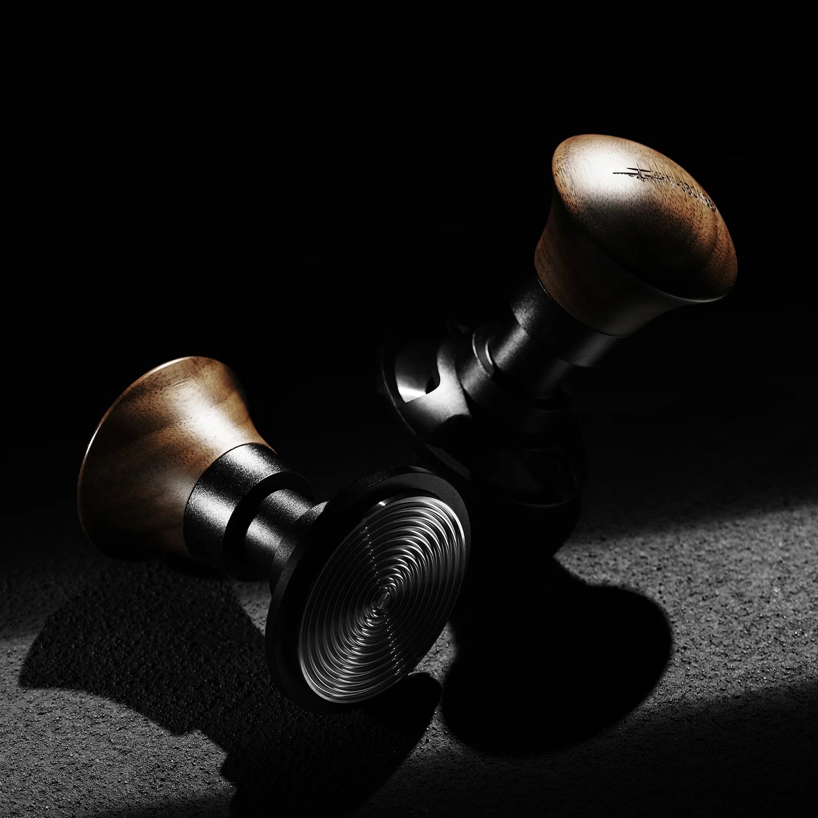 Two MHW-3BOMBER Armor Series Coffee Tamper 2.0 units with wooden handles rest on a dark surface, dramatically lit. Their bases have a shiny spiral pattern and they provide precision with adjustable tamping depth for consistent pressure in every brew.