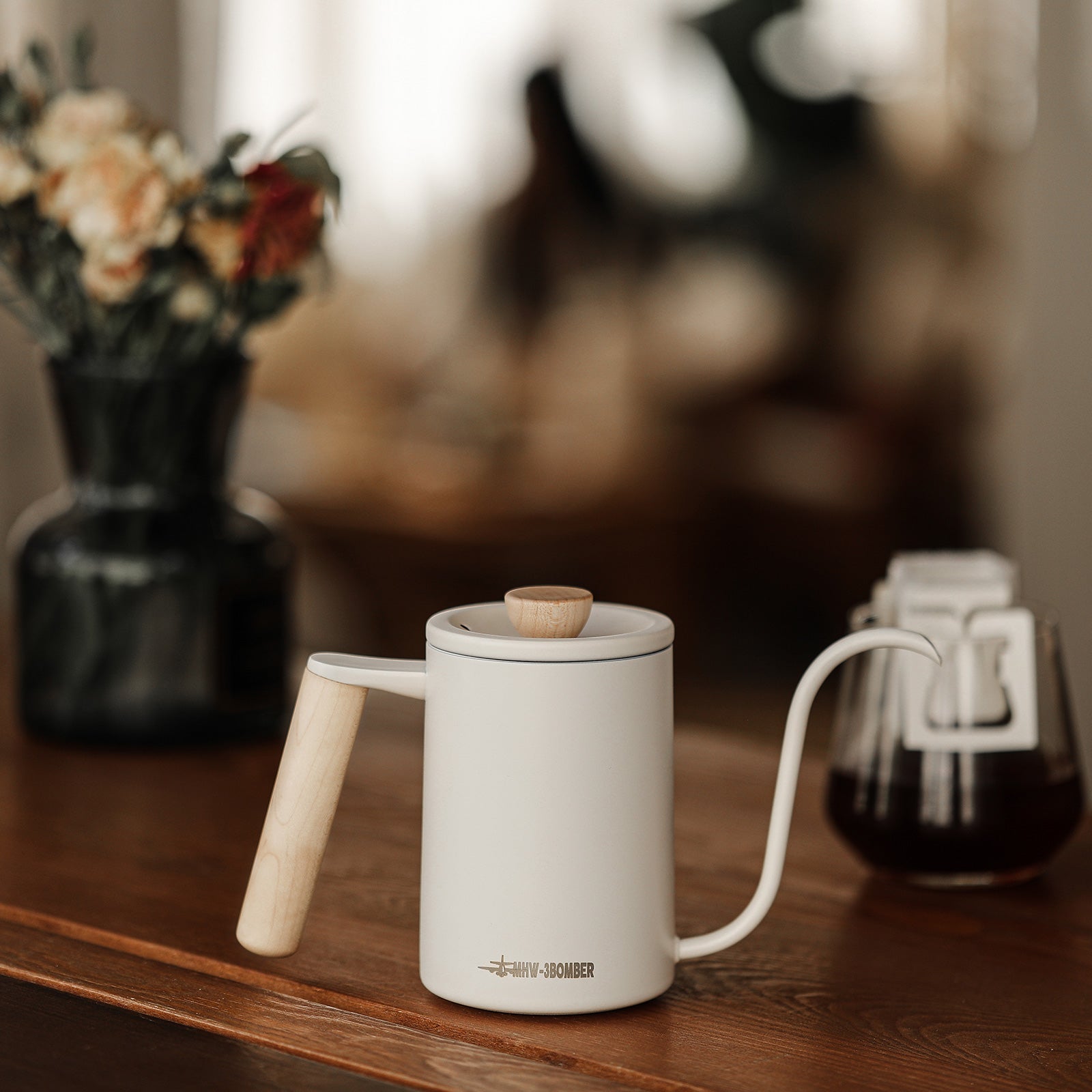 The MHW-3BOMBER Planet Hand Brewing Kettle, a minimalist white gooseneck kettle with a wooden handle and knob, sits on a wooden table. In the background, theres a dark vase with flowers and a glass container. The setting is cozy and softly lit.