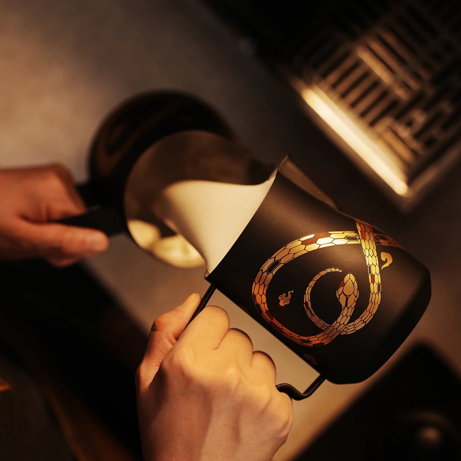 Someone pours milk from the MHW-3BOMBER Turbo Milk Pitcher - Year of the Snake Limited Edition, which features a black design with a golden snake. The scene is warmly lit, creating a cozy atmosphere.