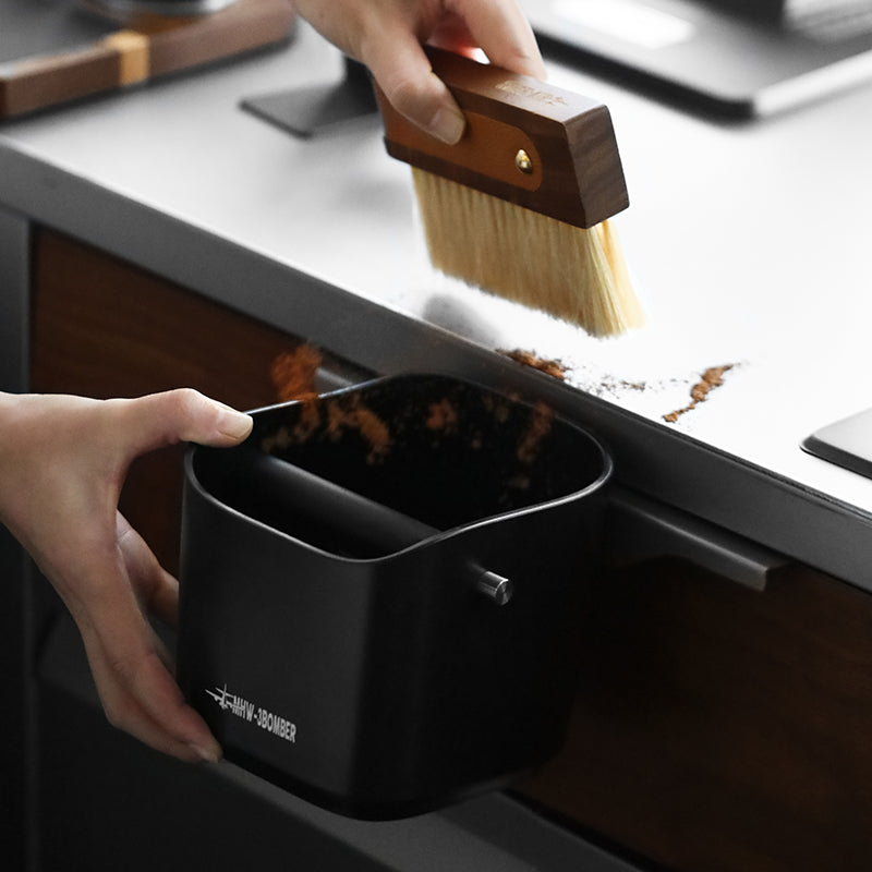 A wooden-handled brush sweeps coffee grounds into a durable black dustpan held at the counters edge, making it easy to clean around your MHW-3BOMBER Coffee Espresso Square Knock Box.