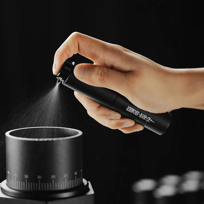 A hand uses a sleek, black MHW-3BOMBER spray bottle from the Ovo Coffee Scale & Spray Bottle Set to create a fine mist over a container. The dark background highlights the delicate mist, ideal for coffee enthusiasts seeking precision comparable to a coffee scale.