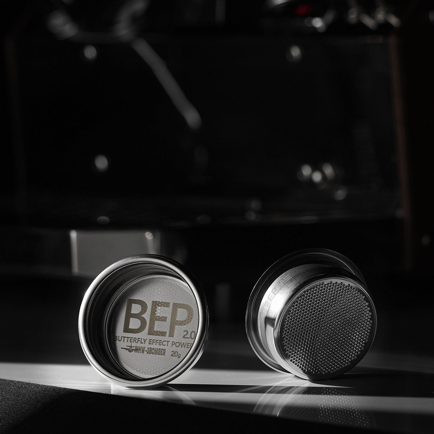Two metallic puck-shaped objects resemble elegant espresso filter baskets on a dark surface. One displays BEP 2.0 BUTTERFLY EFFECT POWER 20g inside, while the other reveals a stainless steel textured surface with dual-layer filtration by MHW-3BOMBERs BEP Butterfly Coffee Filter Basket 2.0 (58.5mm).