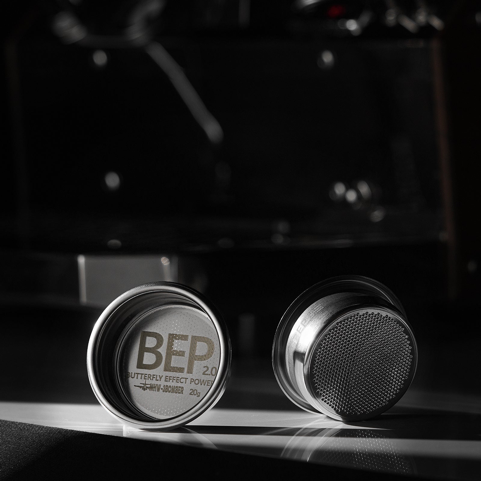 Two metallic puck-shaped objects resemble elegant espresso filter baskets on a dark surface. One displays BEP 2.0 BUTTERFLY EFFECT POWER 20g inside, while the other reveals a stainless steel textured surface with dual-layer filtration by MHW-3BOMBERs BEP Butterfly Coffee Filter Basket 2.0 (58.5mm).
