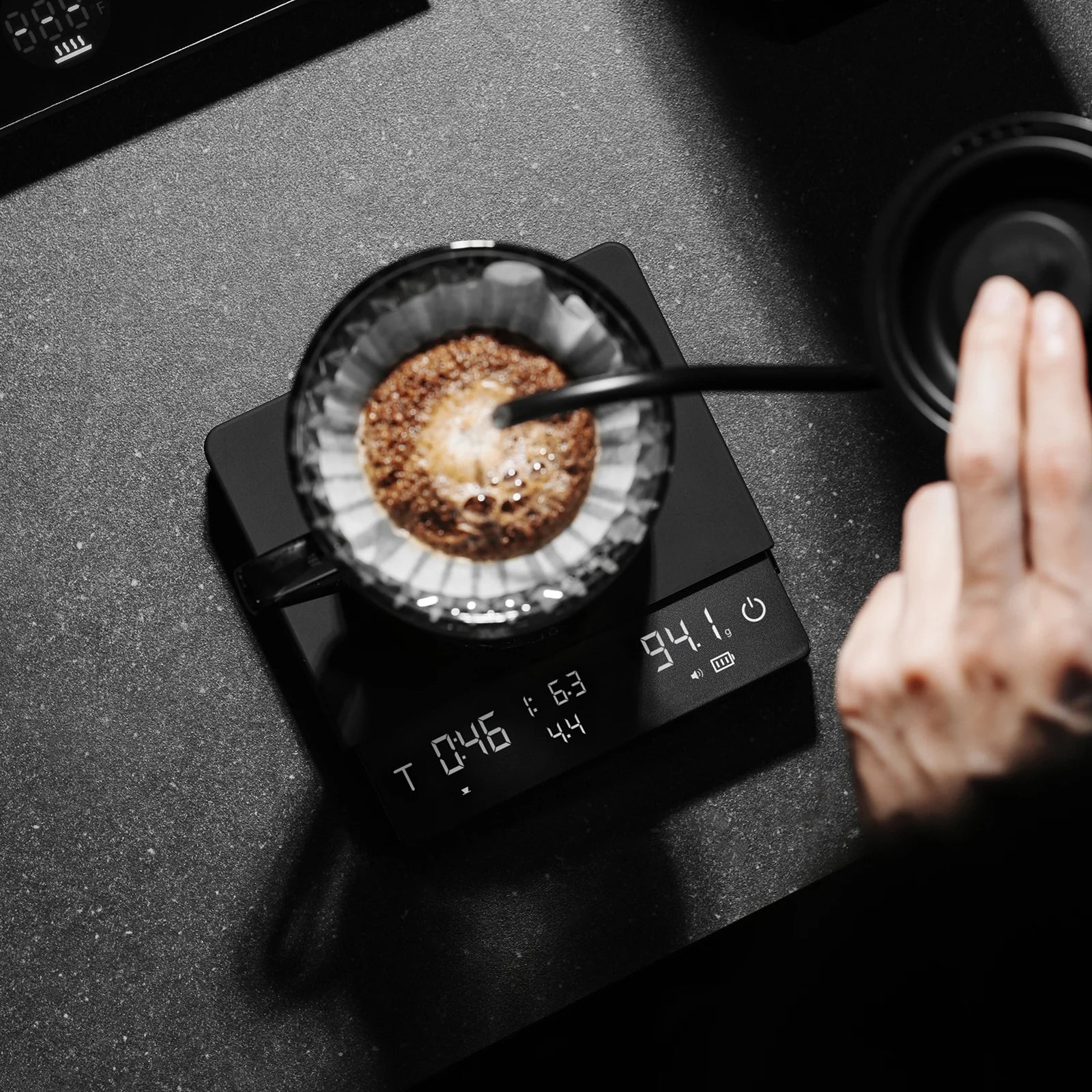 MHW-3BOMBER black Cube Coffee Scale 3.0 Pro Max shown in use, pouring water on a filter. Digital display visible.