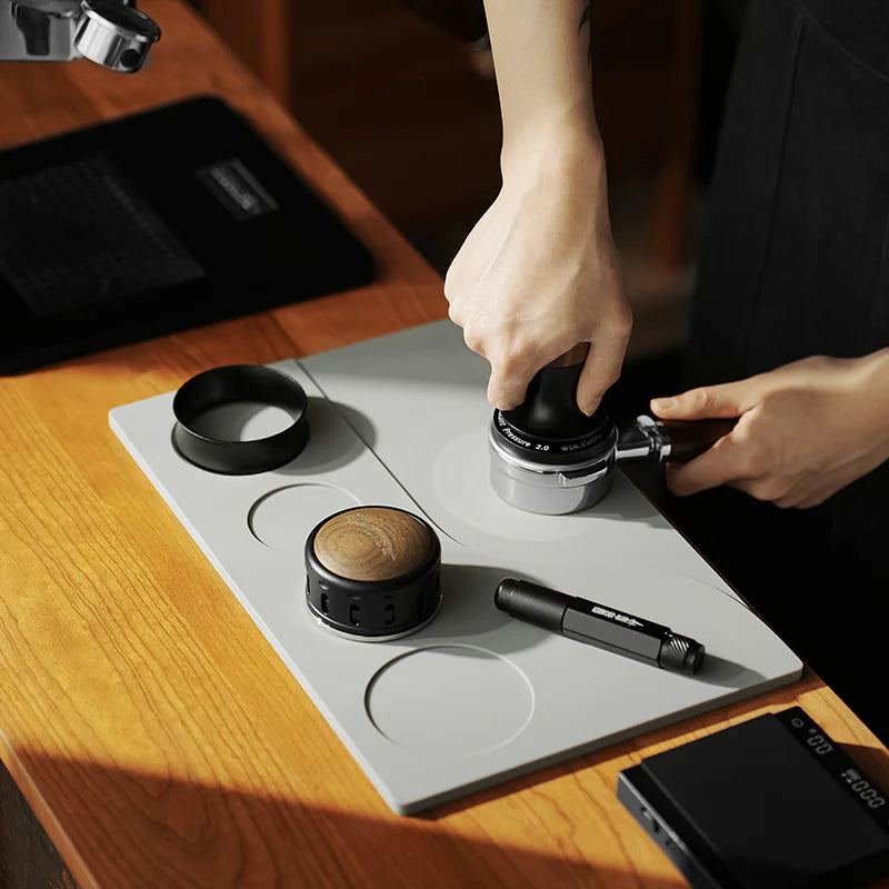 MHW-3BOMBER gray silicone tamping base with portafilter, tamper, and distributor slots on a wood counter. 