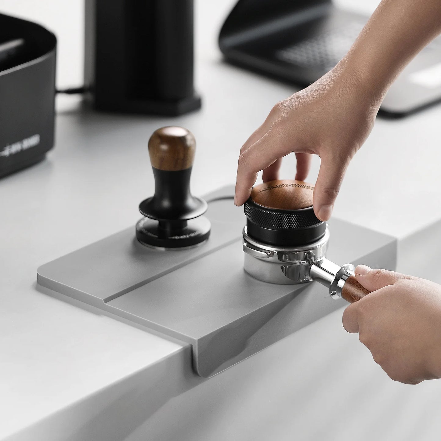MHW-3BOMBER gray silicone coffee tamper mat with two holes, portafilter, and tamper. Used on a kitchen counter.