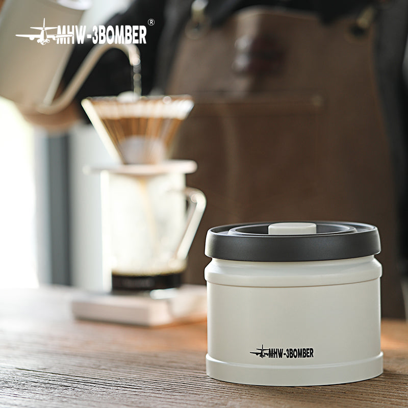 MHW-3BOMBER vacuum coffee canister. Cream color with black lid. Keeps beans fresh. On wooden counter.