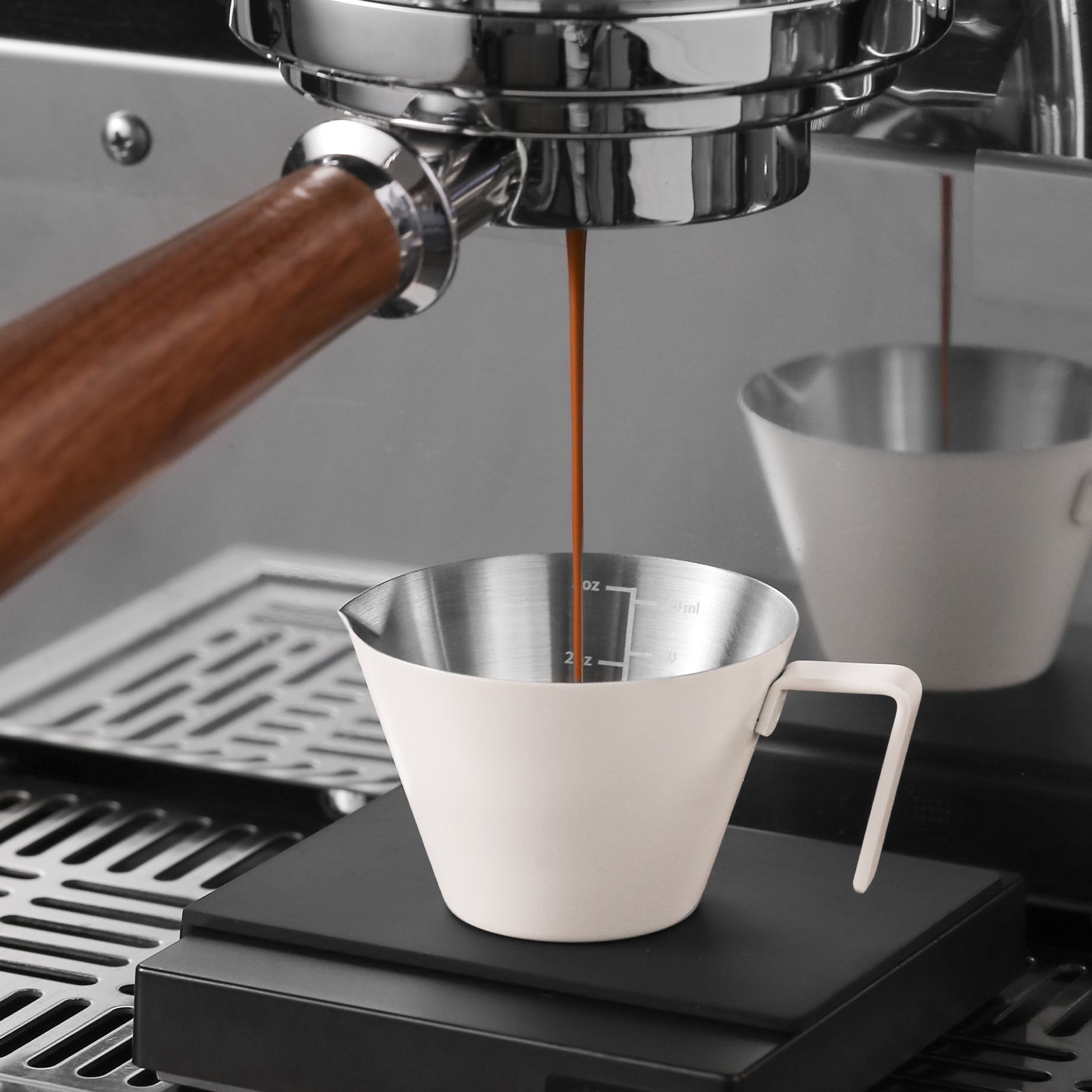 The espresso machine pours coffee into an MHW-3BOMBER Stainless Steel Measuring Cup, its marked interior capturing each rich drop. Coffee brewing takes place on a stainless steel countertop, enhancing the scene with its reflective allure.