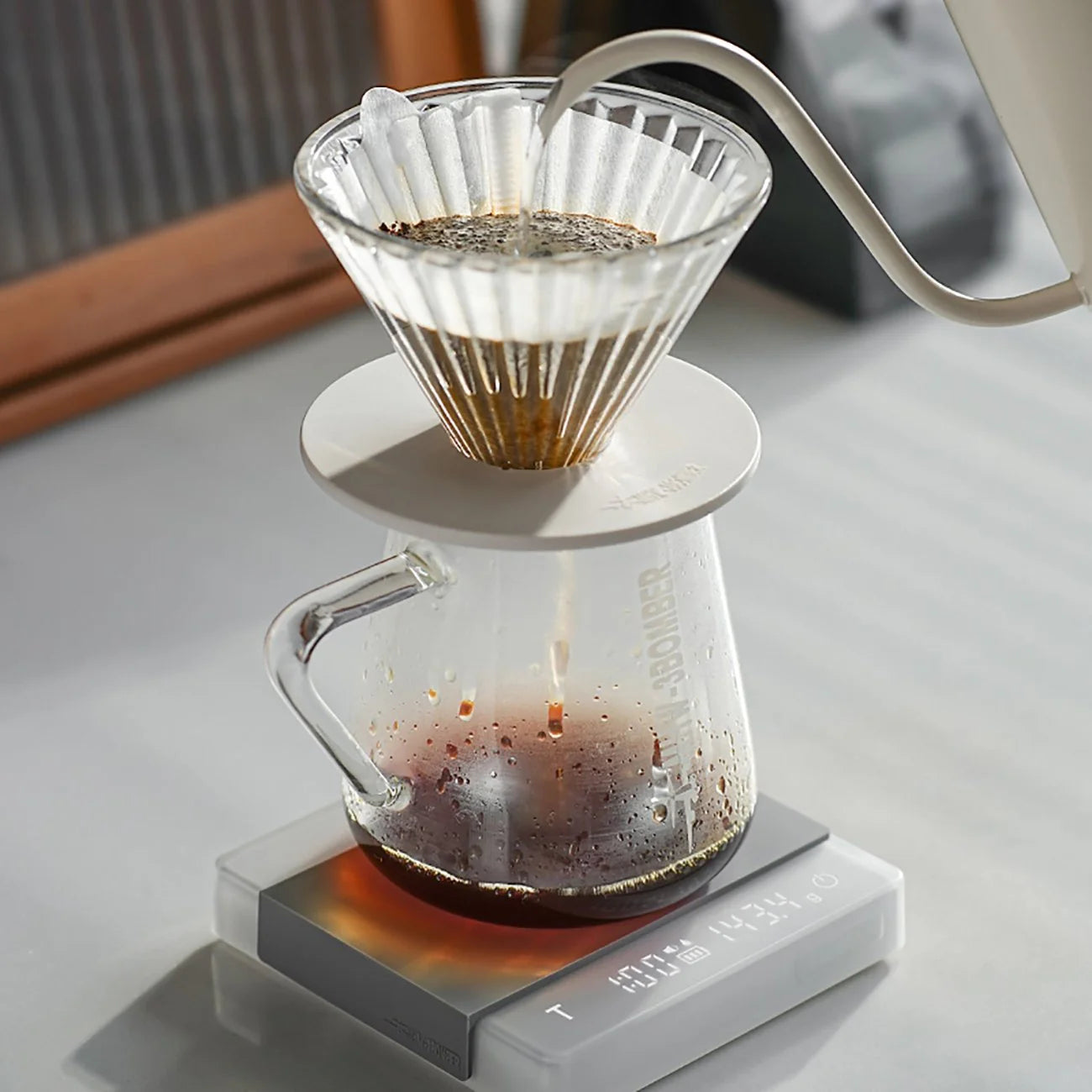 A glass coffee dripper rests on a jug while hot water flows over the grounds. The setup sits on the MHW-3BOMBER Espresso Scale Cube Coffee Scale 2.0 for precise measurements. Natural light enhances the scene as the USB-rechargeable scale ensures a perfect brew below.