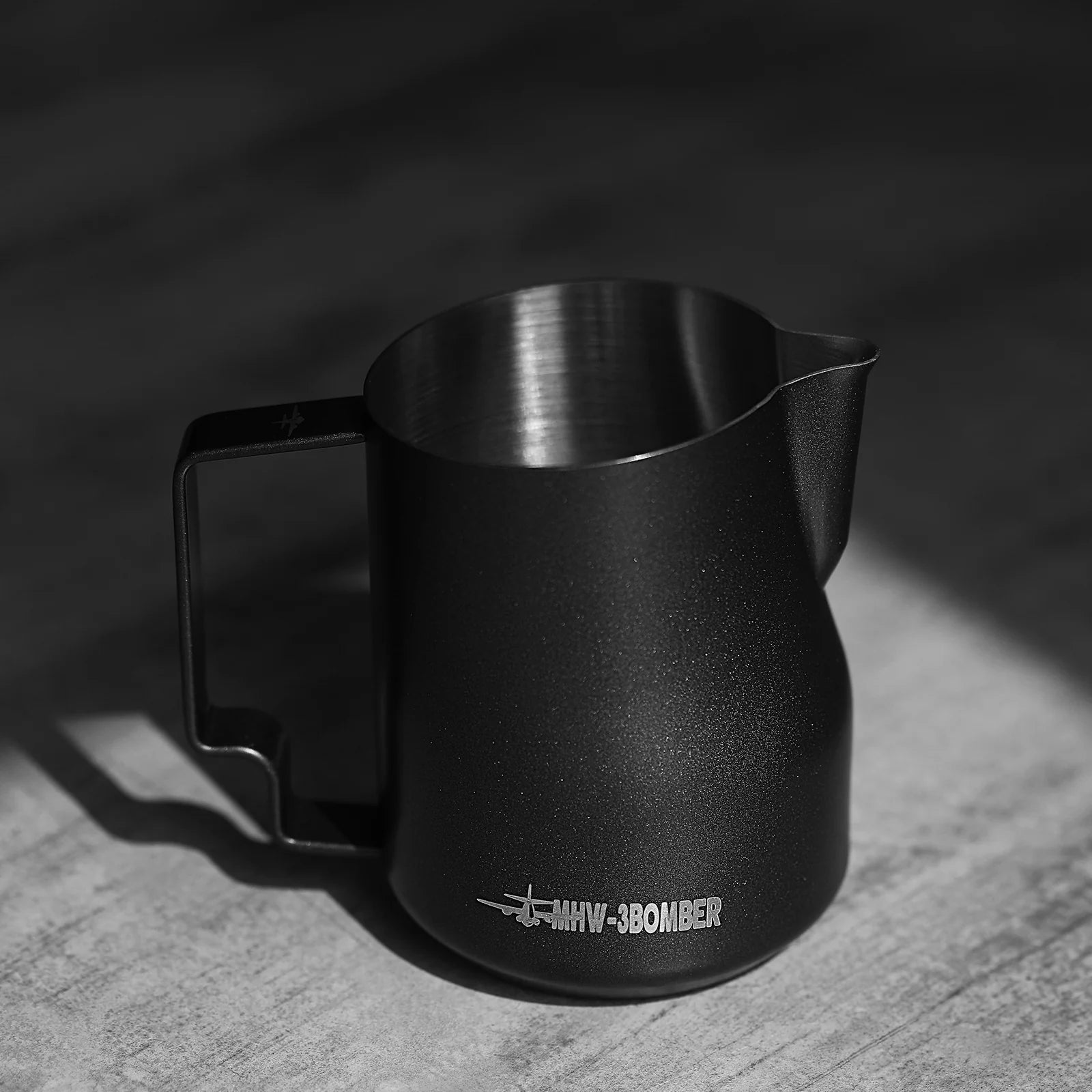 The MHW-3BOMBER Turbo Milk Pitcher Matte Black, with its stainless steel build and sleek handle and spout, sits on a textured gray surface. Perfect for exquisite latte art, it casts subtle shadows as its partially bathed in light.