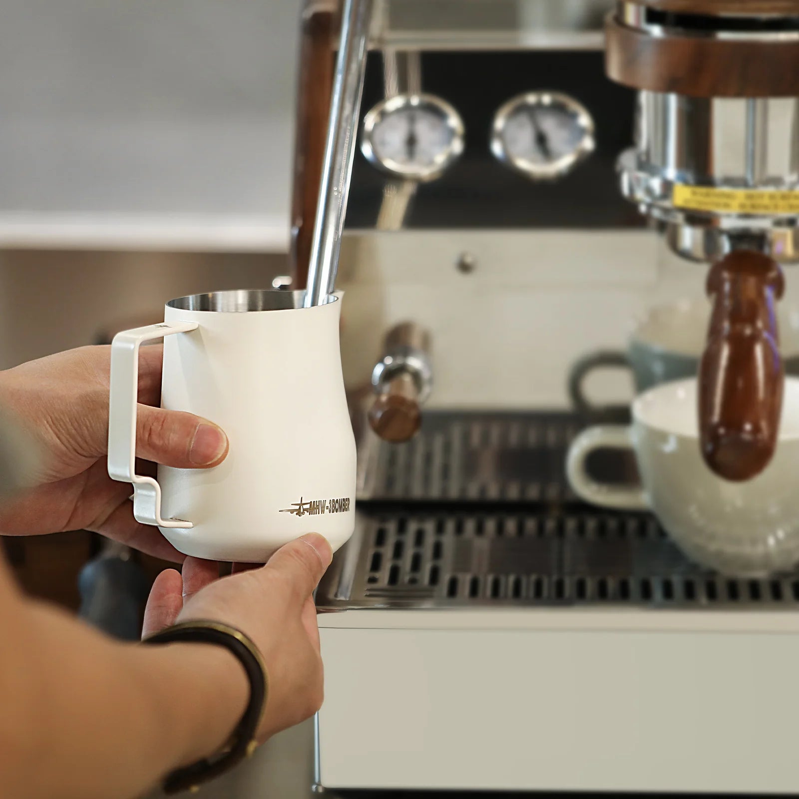 Using the steam wand on a sleek silver espresso machine with dual pressure gauges, someone skillfully froths milk in an MHW-3BOMBER Turbo Milk Pitcher White, while a nearby white mug awaits latte art, complemented by elegant wooden accents.
