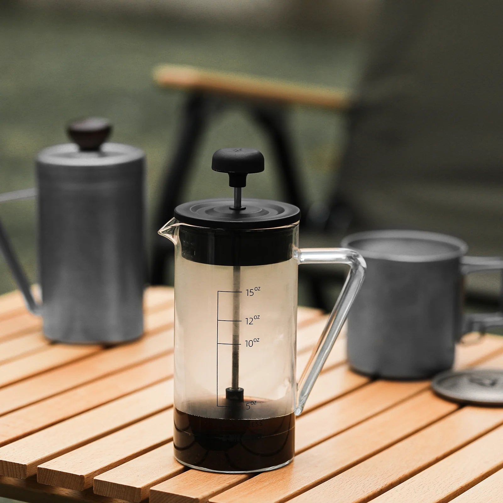 The MHW-3BOMBER Gimme French Press, featuring a sleek design and stainless steel filter, stands proudly on a wooden slatted table outdoors. In the blurred background, two metal mugs and a cylindrical container complete the serene coffee scene.