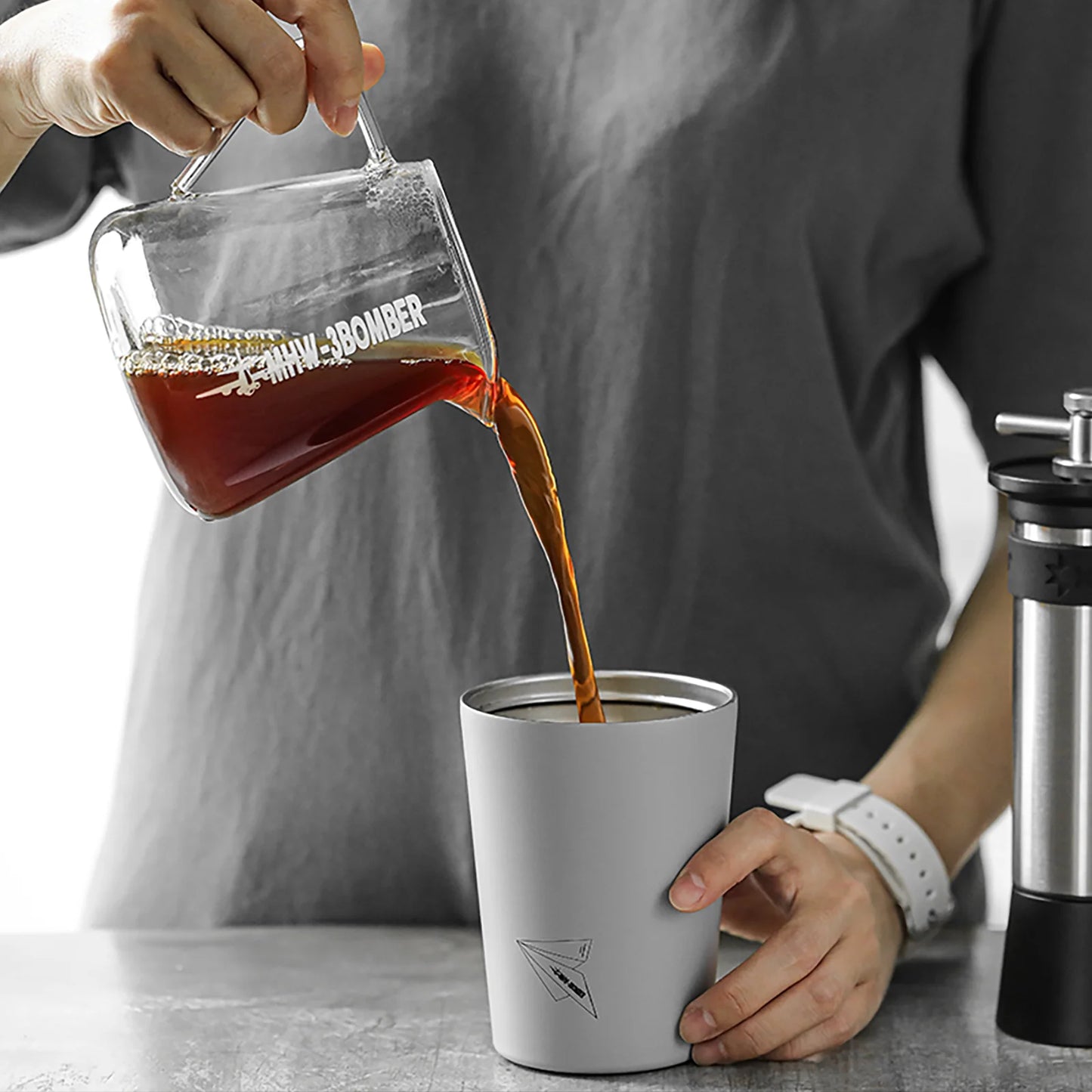 A person in a gray shirt and white watch pours coffee from an MHW-3BOMBER Elf Coffee Server into a gray travel mug with a paper airplane design. This drip-free scene unfolds on a countertop with a coffee grinder nearby.