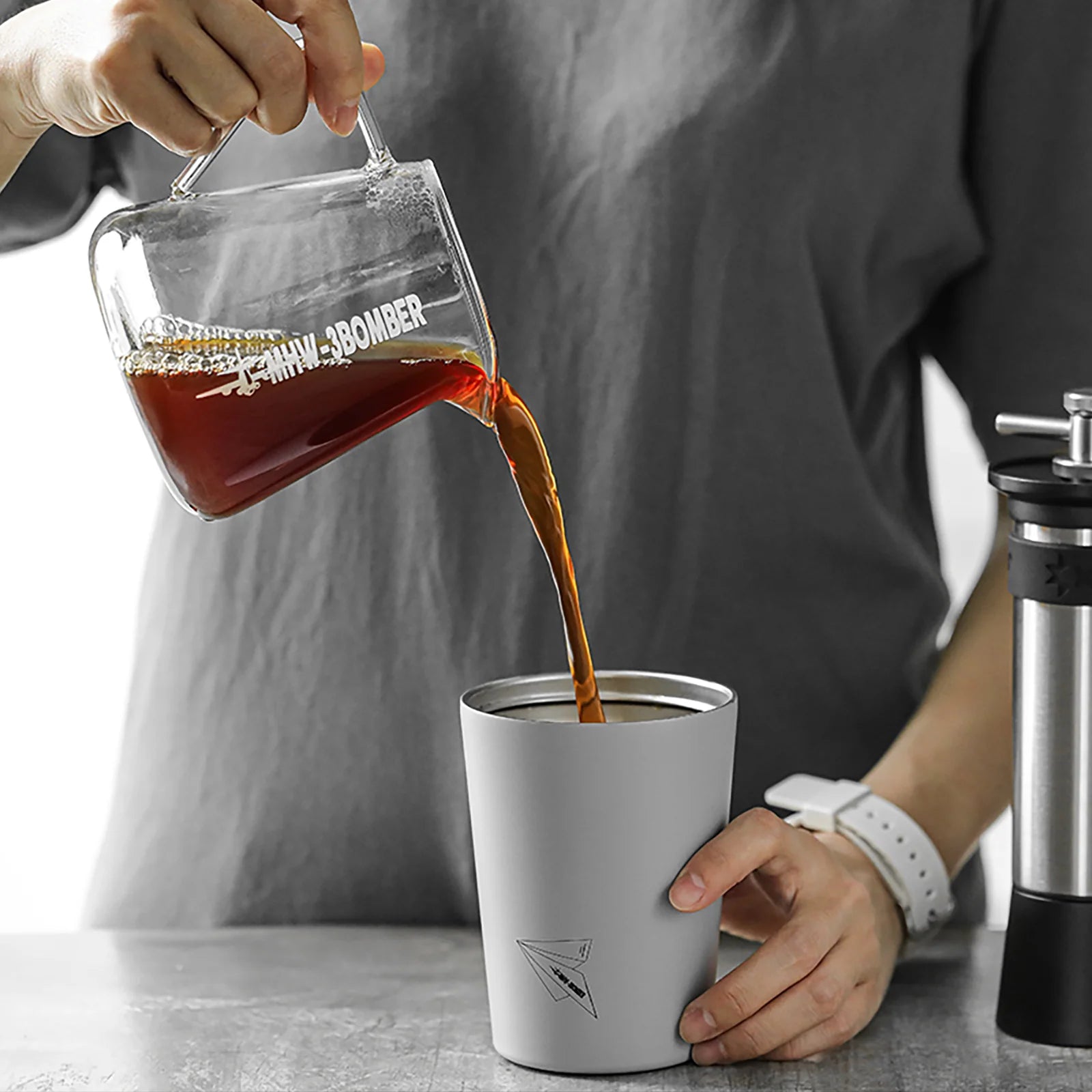 A person in a gray shirt and white watch pours coffee from an MHW-3BOMBER Elf Coffee Server into a gray travel mug with a paper airplane design. This drip-free scene unfolds on a countertop with a coffee grinder nearby.