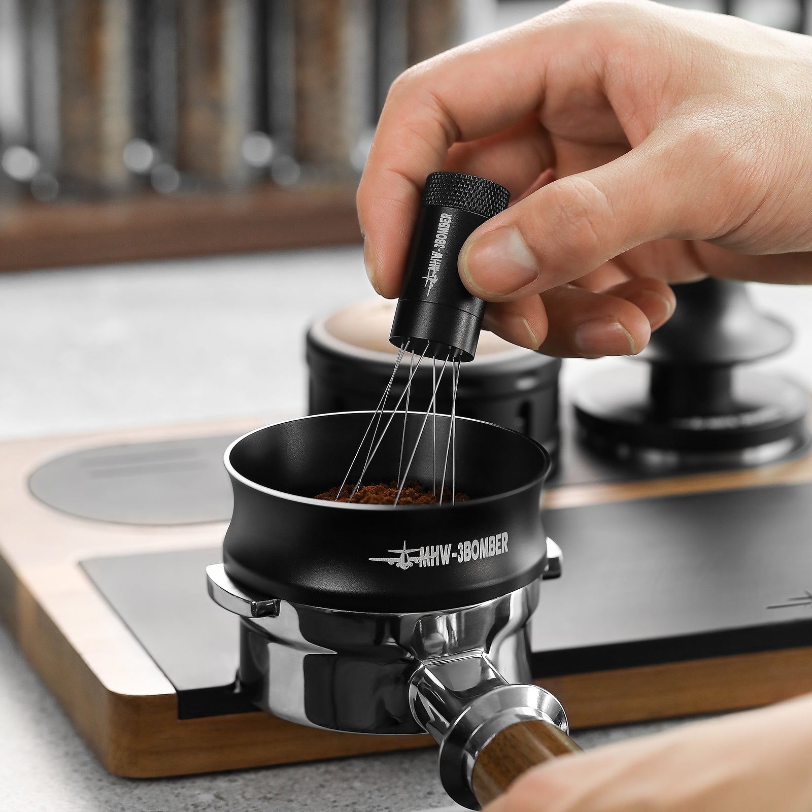 A hand uses a black tool with thin metal prongs to distribute coffee grounds in a portafilter basket, marked MHW-3BOMBER. A tamper, wooden board, and MHW-3BOMBER Espresso Magnetic Coffee Dosing Funnel are visible in the background.