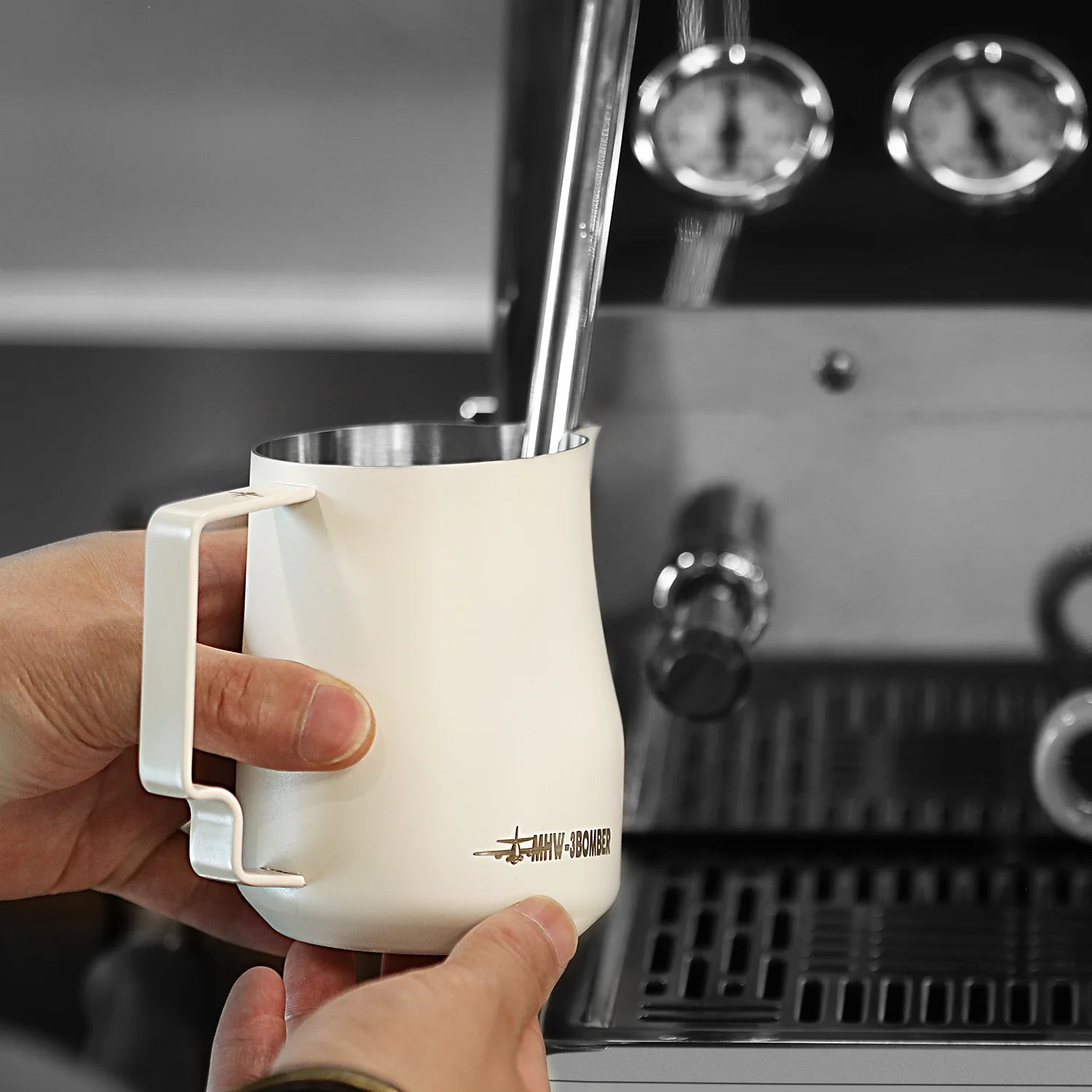 Hands holding an MHW-3BOMBER Turbo Milk Pitcher White, steaming milk with a steam wand on an espresso machine; the focus is on the pitcher and wand, with visible knobs and gauges in the background, ready to create exquisite latte art.