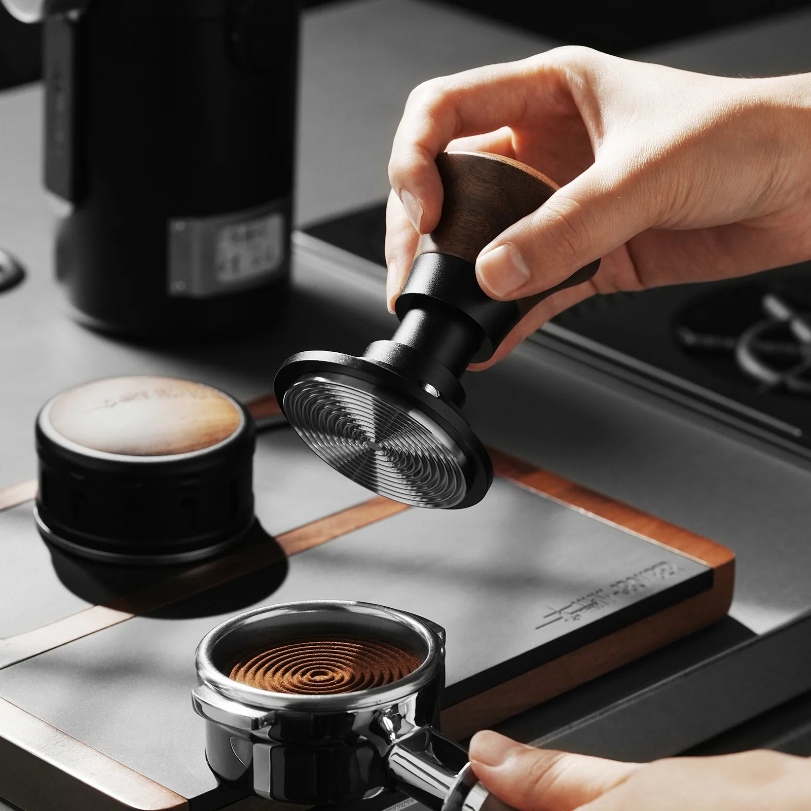 Hands using the MHW-3BOMBER Armor Series Coffee Tamper 2.0, made of metal and wood, to press ground coffee in a portafilter with adjustable depth. A coffee maker with a triple spring design and another tamper are on the gray countertop behind it.