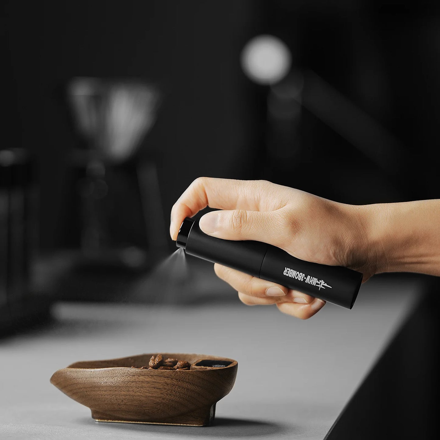A hand sprays liquid from the sleek canister of the MHW-3BOMBER Ovo Coffee Scale & Spray Bottle Set over a wooden bowl of coffee beans, enhancing the brewing process, with a blurred background showcasing a distant coffee dripper.