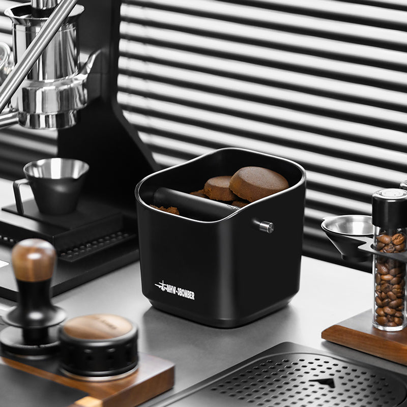 A modern coffee station features the durable MHW-3BOMBER Coffee Espresso Square Knock Box filled with used grounds, easy to clean for convenience. It is surrounded by a coffee machine, portafilter holder, beans, and espresso accessories against a backdrop of horizontal blinds.