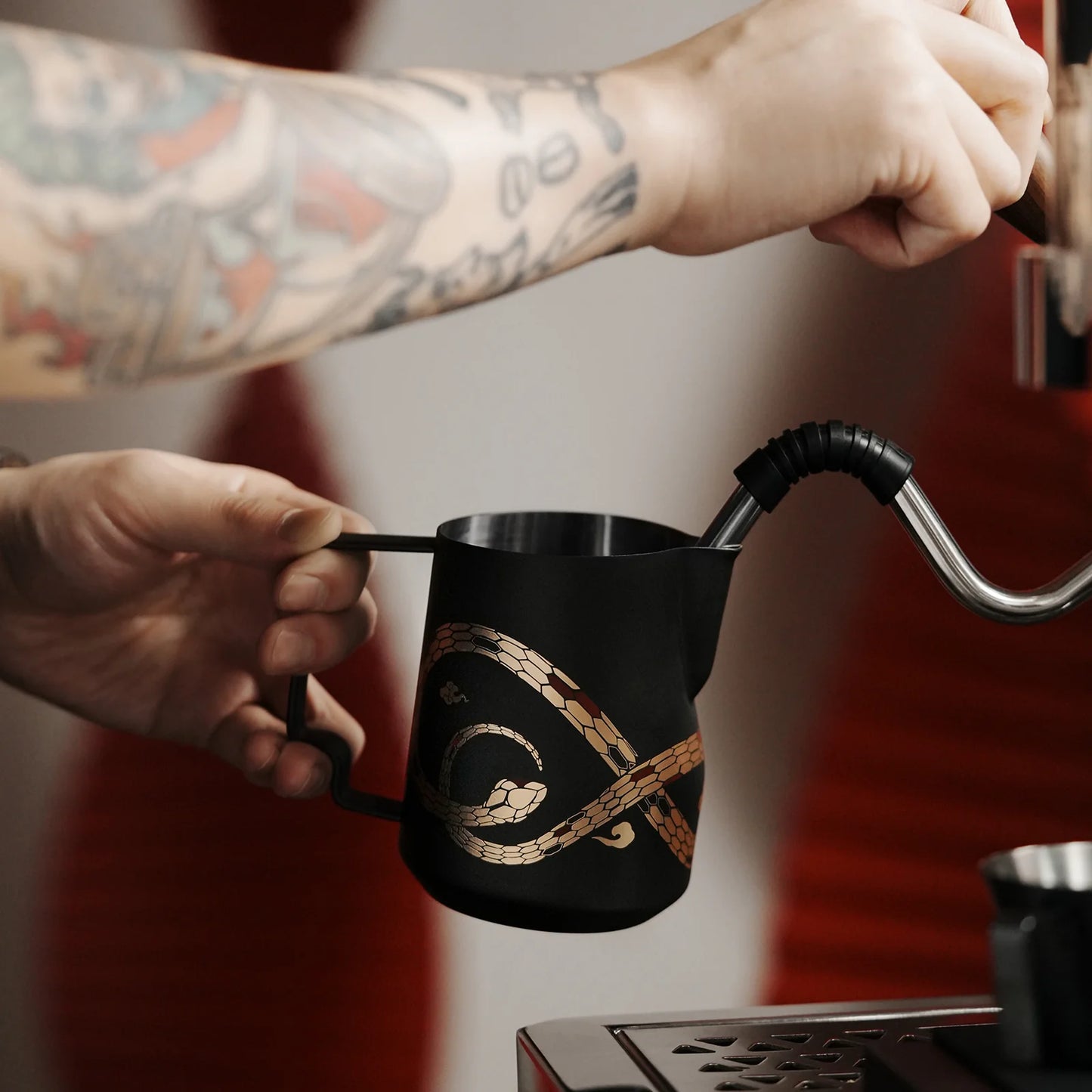 A tattooed individual uses an espresso machines steam wand to froth milk in the MHW-3BOMBER Turbo Milk Pitcher-Year of the Snake Limited Edition. The sleek black pitcher with its gold design stands out against a backdrop of blurred red and white elements, creating a vibrant contrast.
