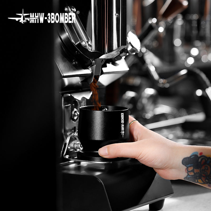 A barista uses the MHW-3BOMBER Espresso Coffee Powder Dosing Funnel under a sleek coffee grinder, expertly catching ground coffee. The MHW-3BOMBER brand shines as the blurred background emphasizes their brewing mastery.