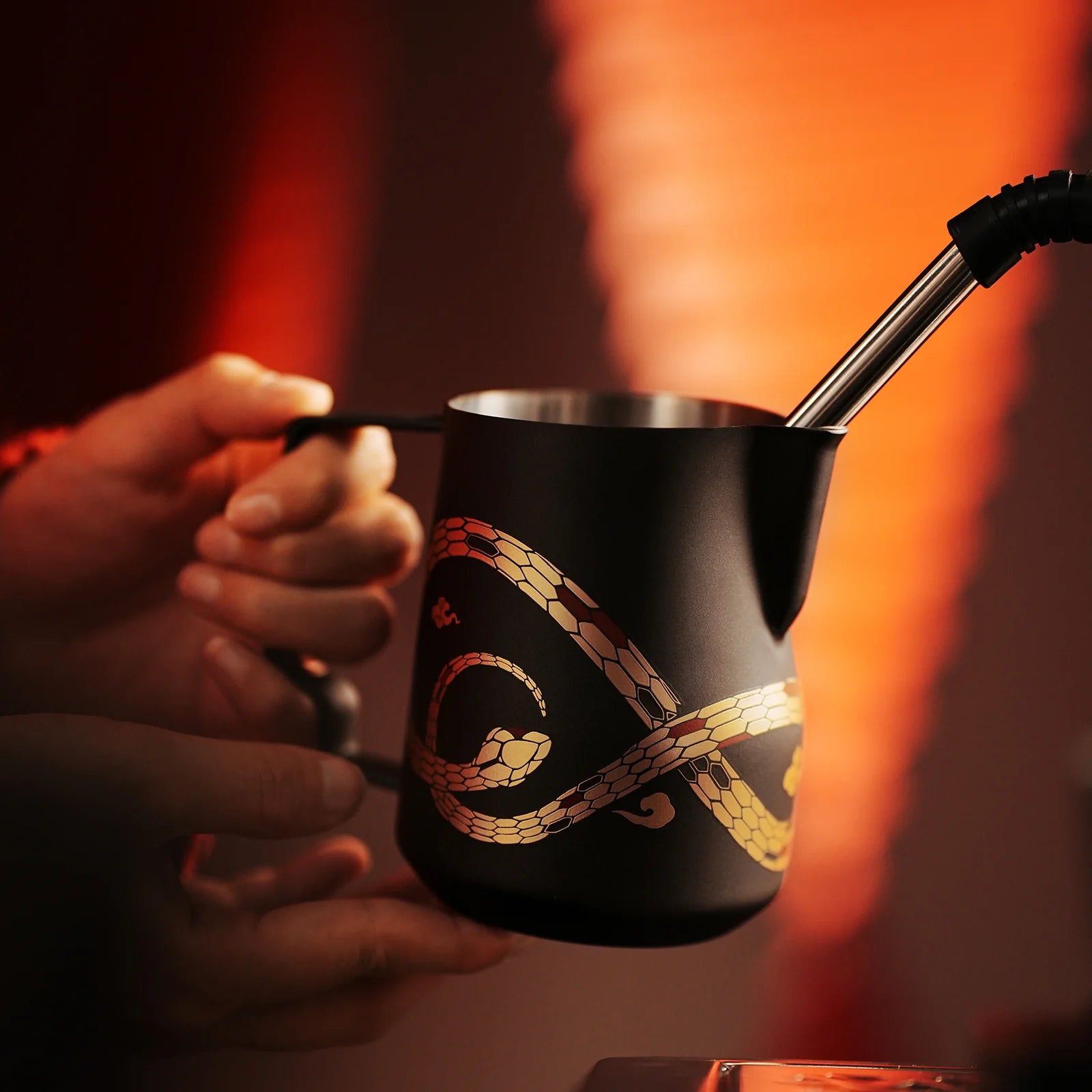 Hands holding the MHW-3BOMBER Turbo Milk Pitcher-Year of the Snake Limited Edition, a black pitcher with a silver spout crafted from Austenitic 304 Stainless Steel and featuring a snake pattern. The softly illuminated warm-toned background creates a cozy atmosphere.