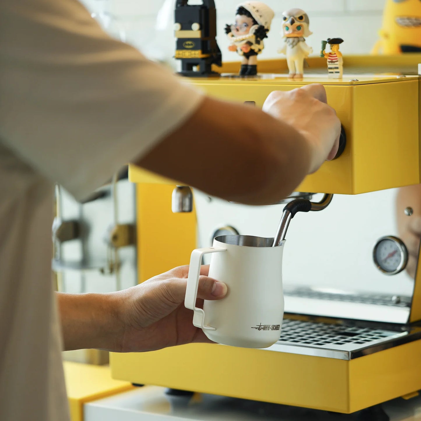 Using a MHW-3BOMBER espresso machine adorned with small figurines, a person expertly froths milk in the MHW-3BOMBER Turbo Milk Pitcher White, setting the stage for exquisite latte art.