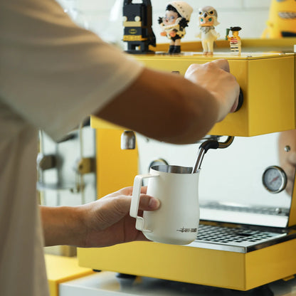 Using a MHW-3BOMBER espresso machine adorned with small figurines, a person expertly froths milk in the MHW-3BOMBER Turbo Milk Pitcher White, setting the stage for exquisite latte art.