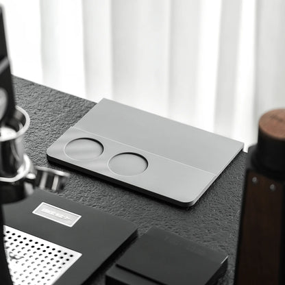 MHW-3BOMBER coffee tamper mat. Silicone pad, silver with two holes. On a counter.