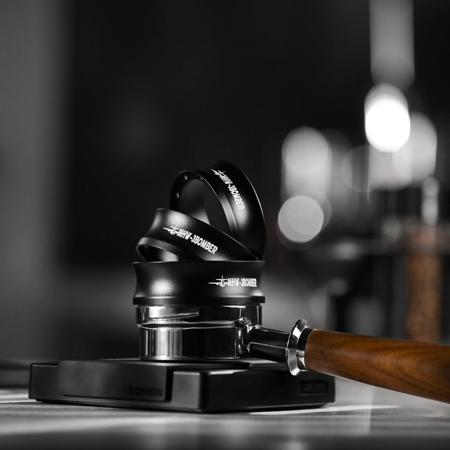 A wooden-handled espresso tamper with a polished base sits on the counter next to stacked MHW-3BOMBER Espresso Magnetic Coffee Dosing Funnels. The softly blurred background highlights these coffee-making tools.