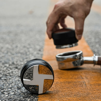 MHW-3BOMBER coffee distributor, black with silver cross design. Coffee tamper on a textured surface.