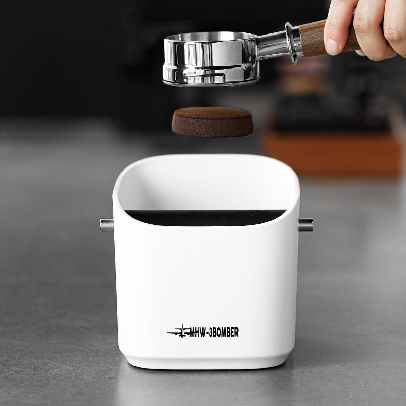 A MHW-3BOMBER Coffee Espresso Square Knock Box rests on a metallic surface, featuring a sturdy metal bar for easy cleaning. A hand with a portafilter hovers above, poised to effortlessly discard the used coffee puck.