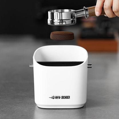 A MHW-3BOMBER Coffee Espresso Square Knock Box rests on a metallic surface, featuring a sturdy metal bar for easy cleaning. A hand with a portafilter hovers above, poised to effortlessly discard the used coffee puck.