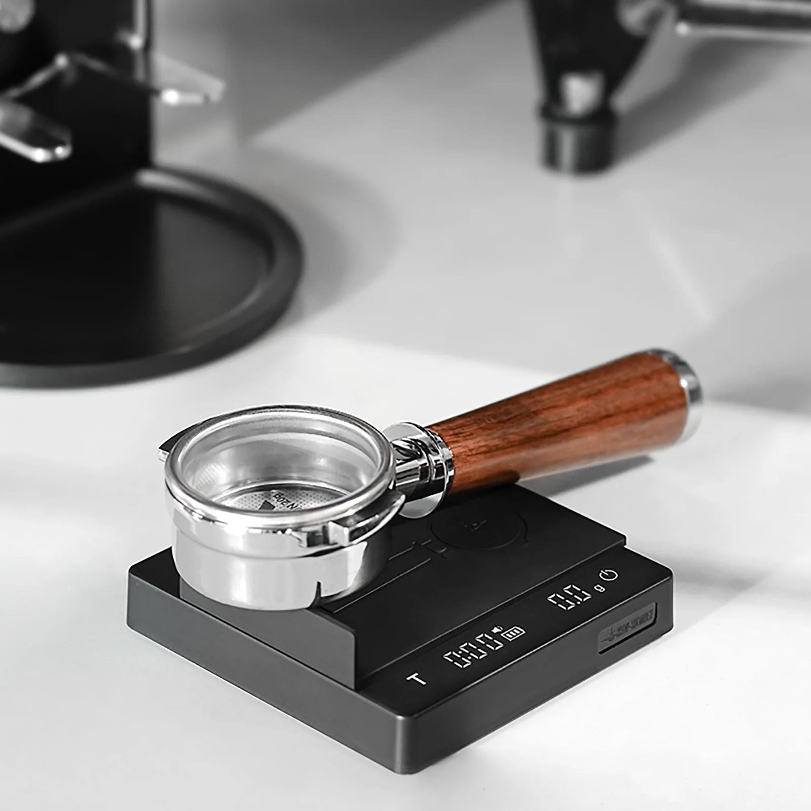A portafilter with a wooden handle rests on an MHW-3BOMBER Espresso Scale Cube Coffee Scale 2.0, USB rechargeable, displaying numbers on a white countertop as part of the coffee-making setup, with an espresso machine visible in the background.