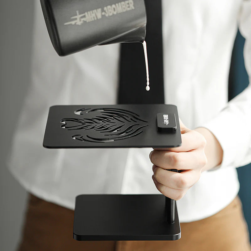 MHW-3BOMBER coffee scale stand in black; barista pouring milk. Height-adjustable, waterproof, silicone base. 