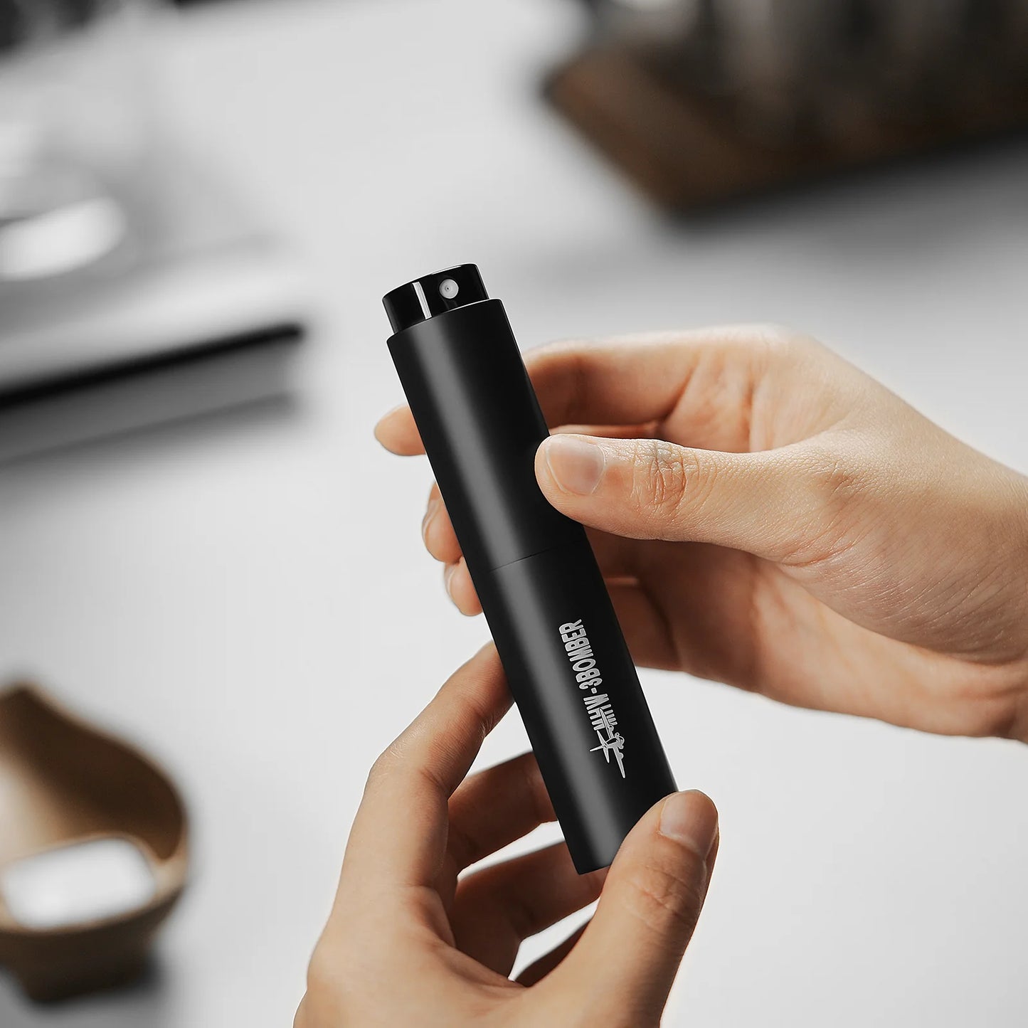 A person is holding a sleek, black cylindrical spray bottle from the MHW-3BOMBER Ovo Coffee Scale & Spray Bottle Set. It features white text, contrasting against a blurred background of a wooden tray and small box, reminiscent of precision youd find in an espresso scale.